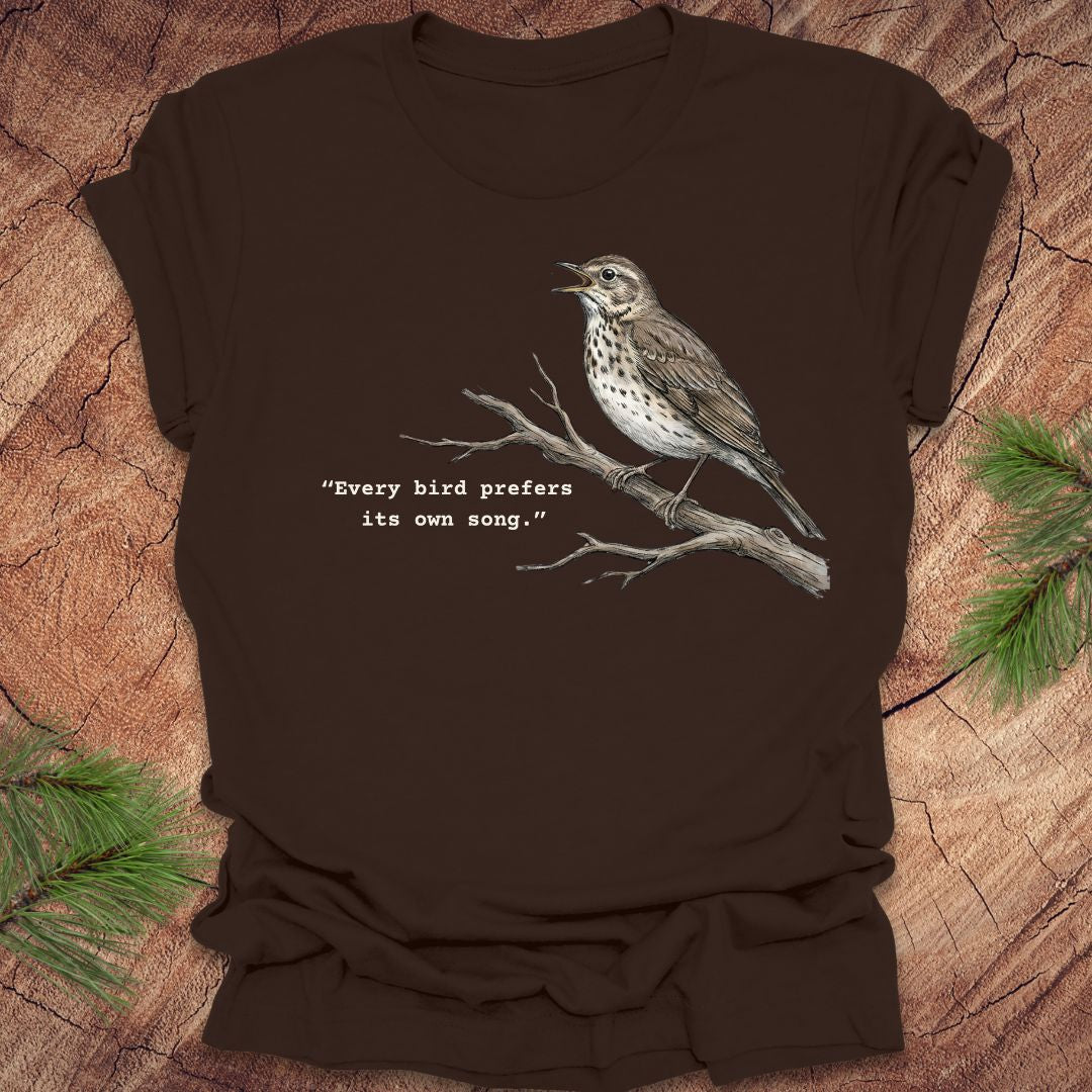 Brown t-shirt with a songbird on a branch and text, 'every bird prefers its own song,' on a wooden background.