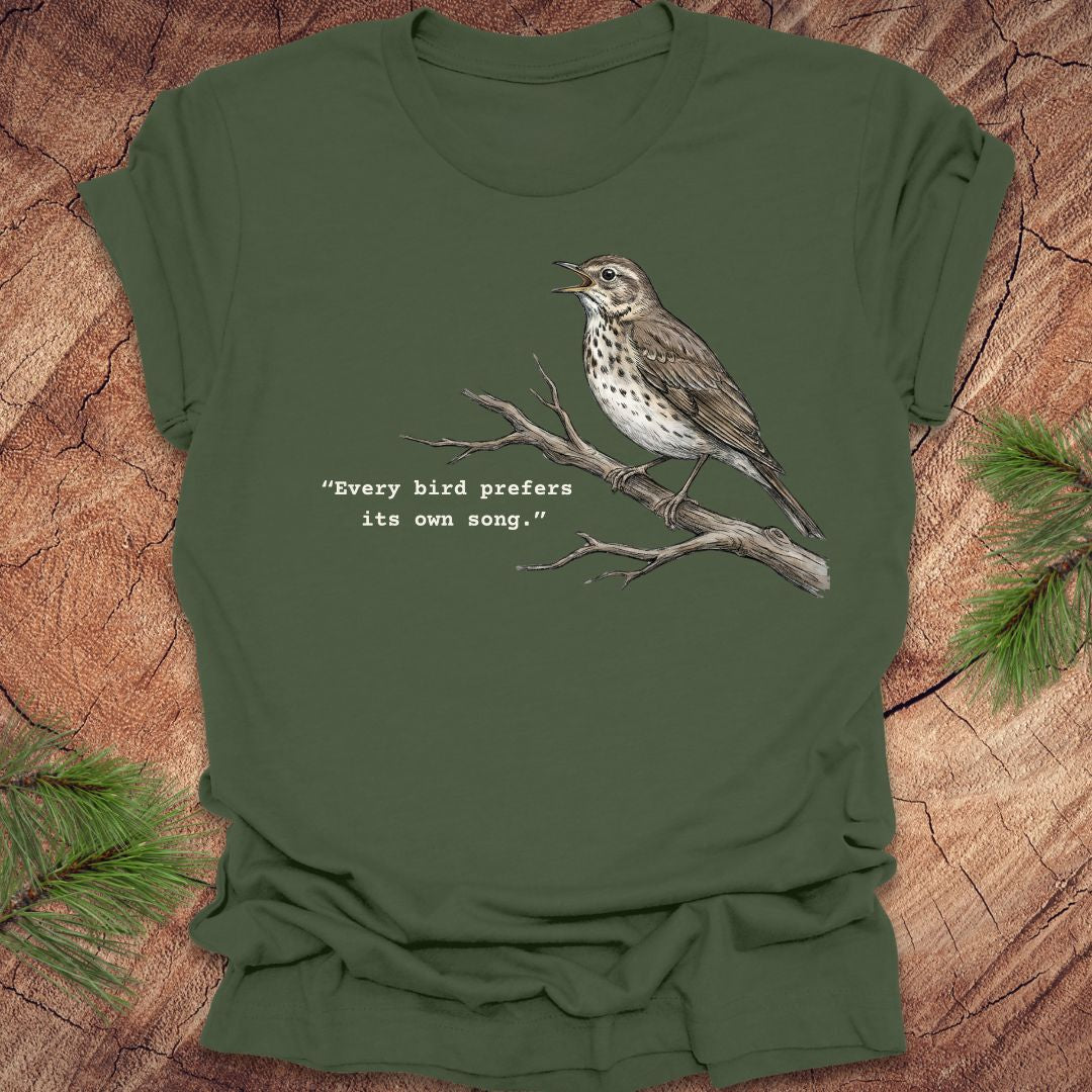 Green t-shirt with a songbird on a branch and text, 'every bird prefers its own song,' on a wooden background.