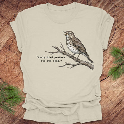 Beige t-shirt with a songbird on a branch and text, 'every bird prefers its own song,' on a wooden background.