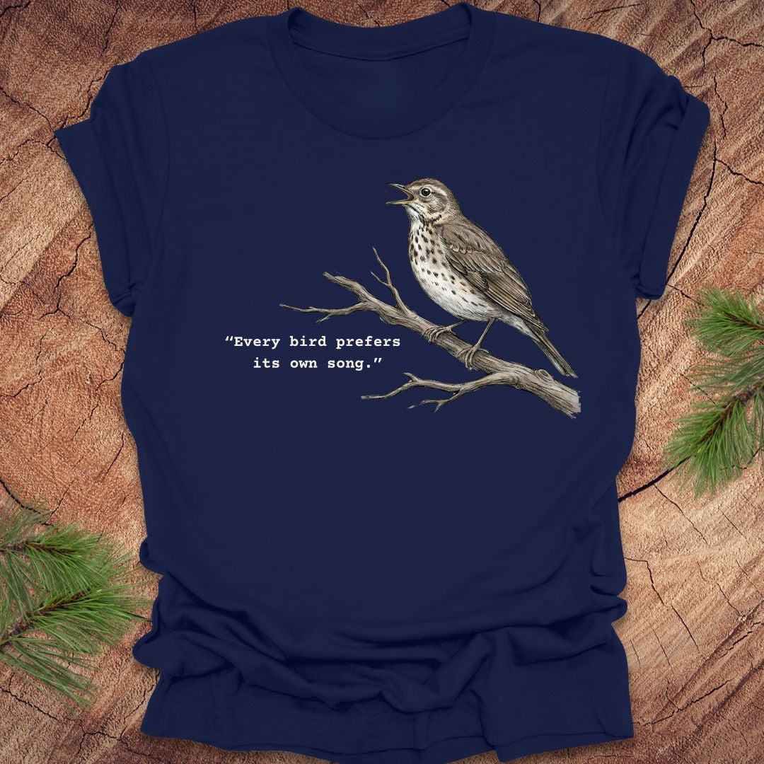 Navy blue t-shirt with a songbird on a branch and text, 'every bird prefers its own song,' on a wooden background.
