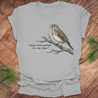 Silver t-shirt with a songbird on a branch and text, 'every bird prefers its own song,' on a wooden background.