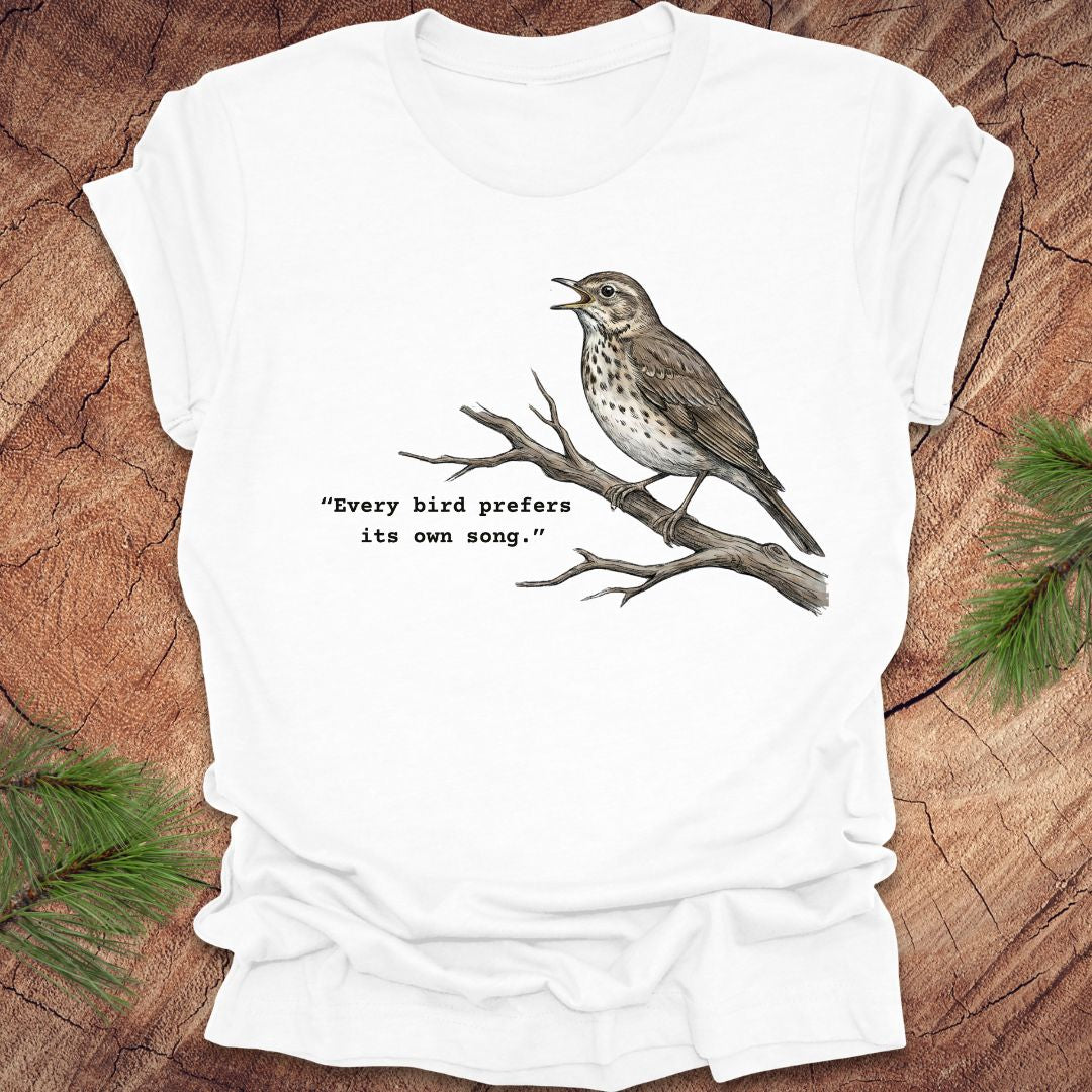 White t-shirt with a songbird on a branch and text, 'every bird prefers its own song,' on a wooden background.