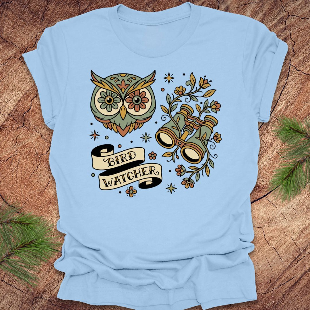 Baby blue t-shirt with owl and binoculars design, with text 'bird watcher' on a wooden surface