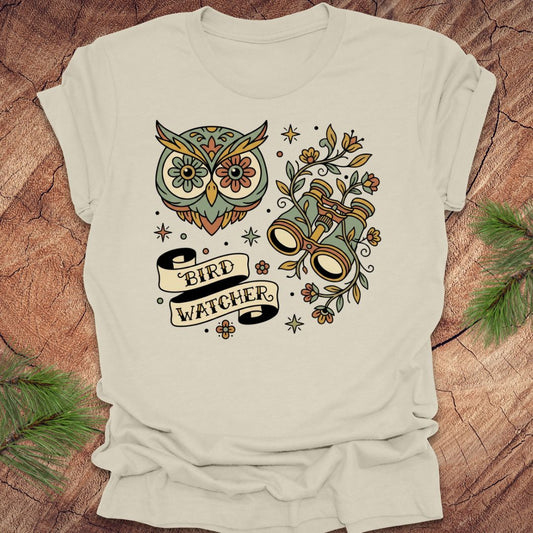 Beige t-shirt with owl and binoculars design, with text 'bird watcher' on a wooden surface