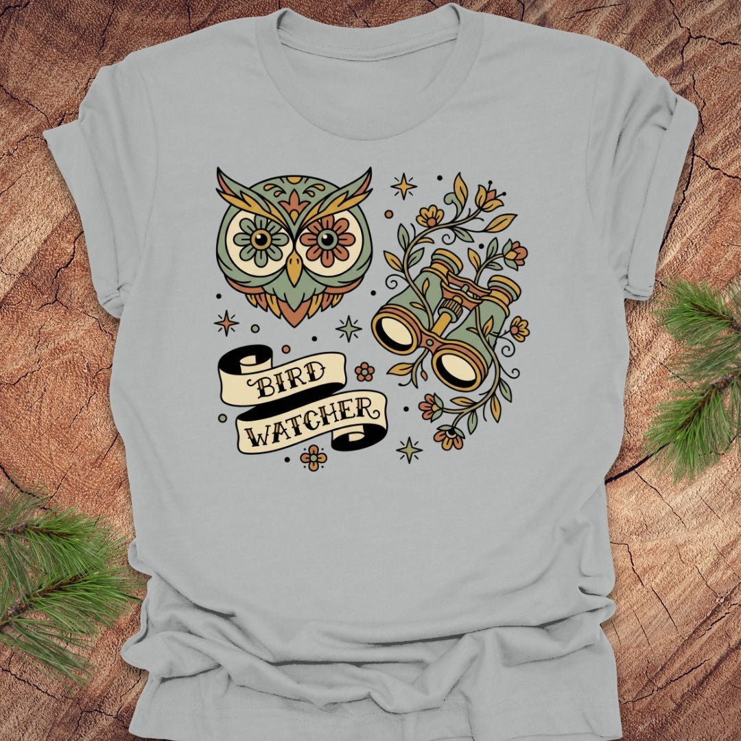 Gray t-shirt with owl and binoculars design, with text 'bird watcher' on a wooden surface