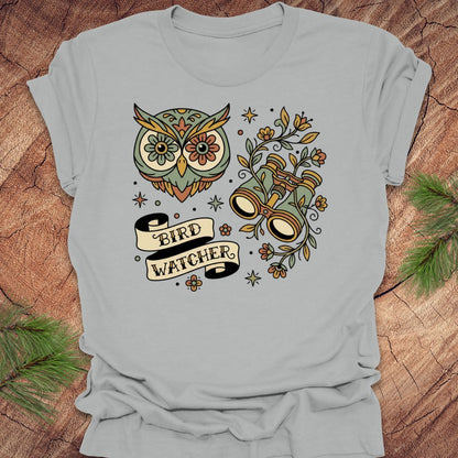 Gray t-shirt with owl and binoculars design, with text 'bird watcher' on a wooden surface