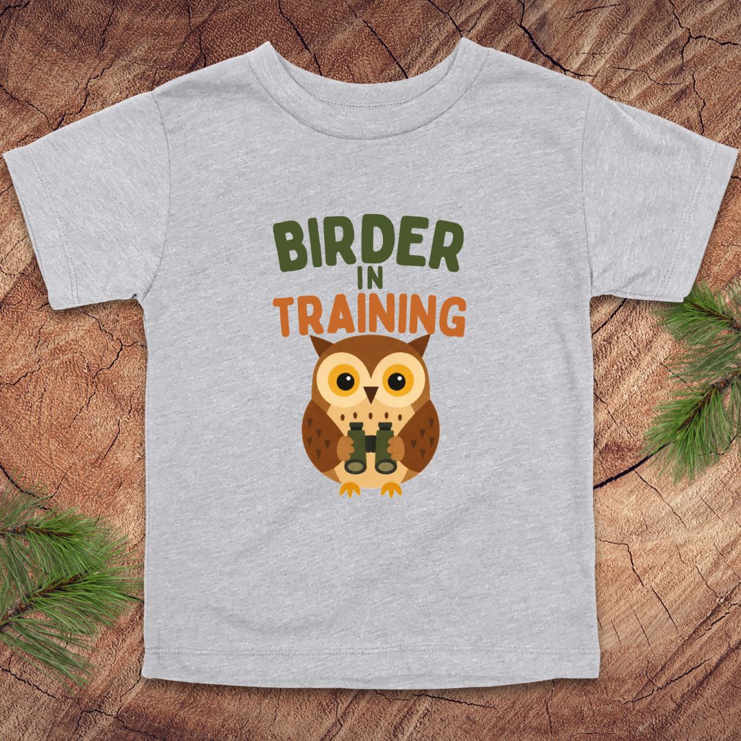 Gray t-shirt with an owl graphic and 'Birder in Training' text on a wooden surface.