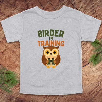 Gray t-shirt with an owl graphic and 'Birder in Training' text on a wooden surface.