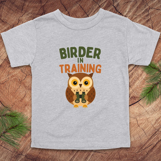 Gray t-shirt with an owl graphic and 'Birder in Training' text on a wooden surface.
