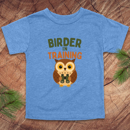 Blue t-shirt with an owl graphic and 'Birder in Training' text on a wooden surface.
