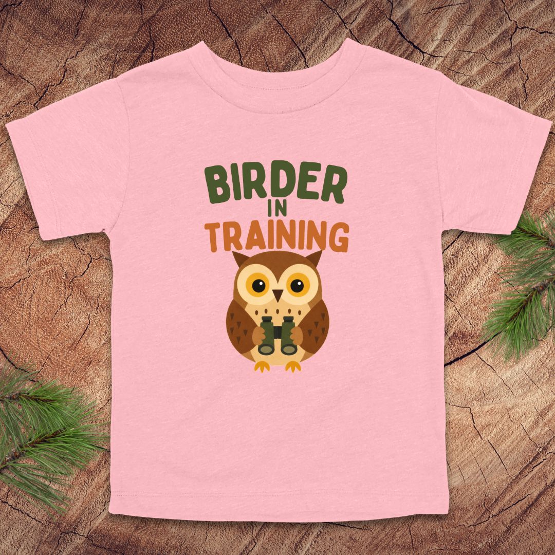 Pink t-shirt with owl graphic and 'Birder in Training' text on a wooden surface.