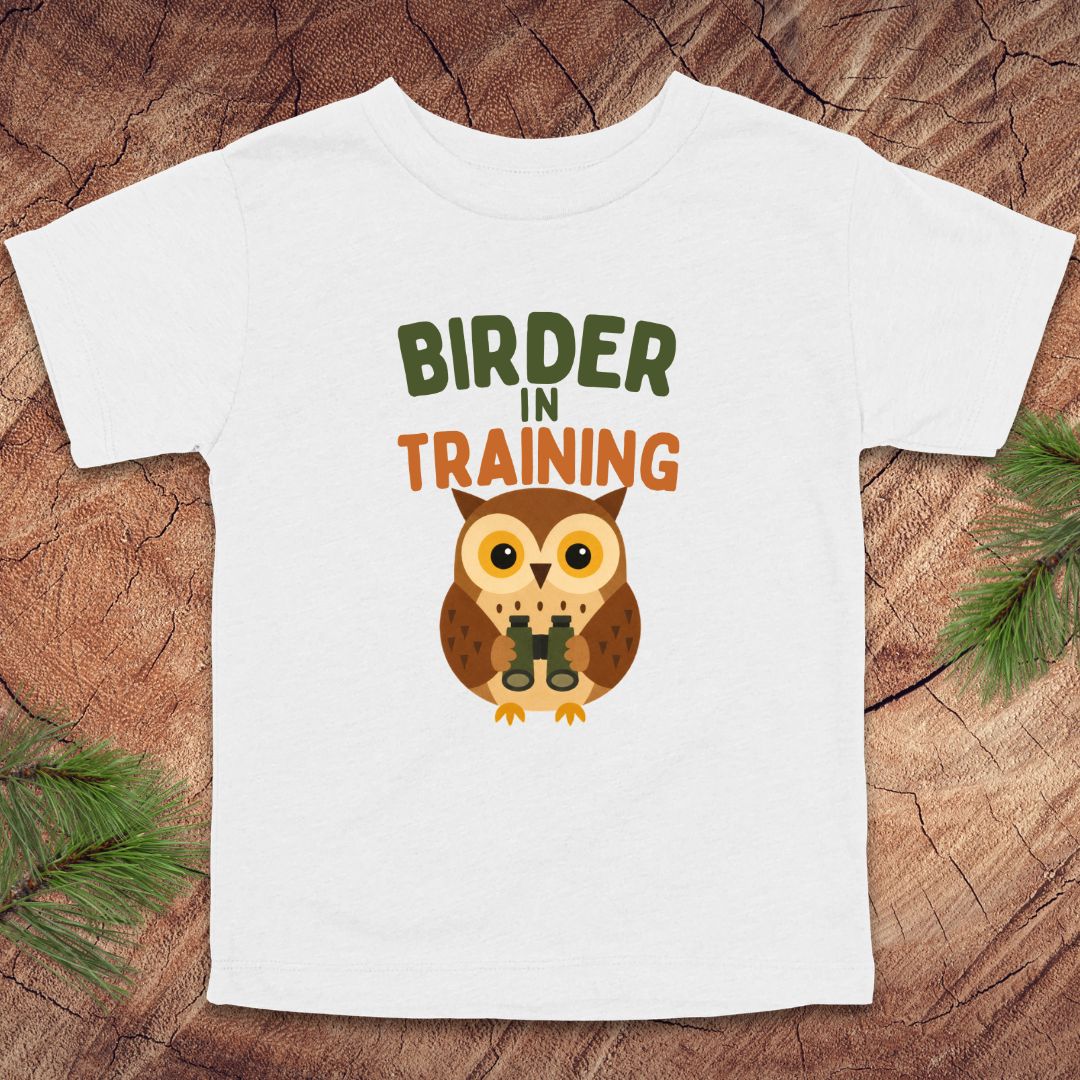 White t-shirt with an owl graphic and 'Birder in Training' text on a wooden surface.