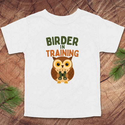 White t-shirt with an owl graphic and 'Birder in Training' text on a wooden surface.