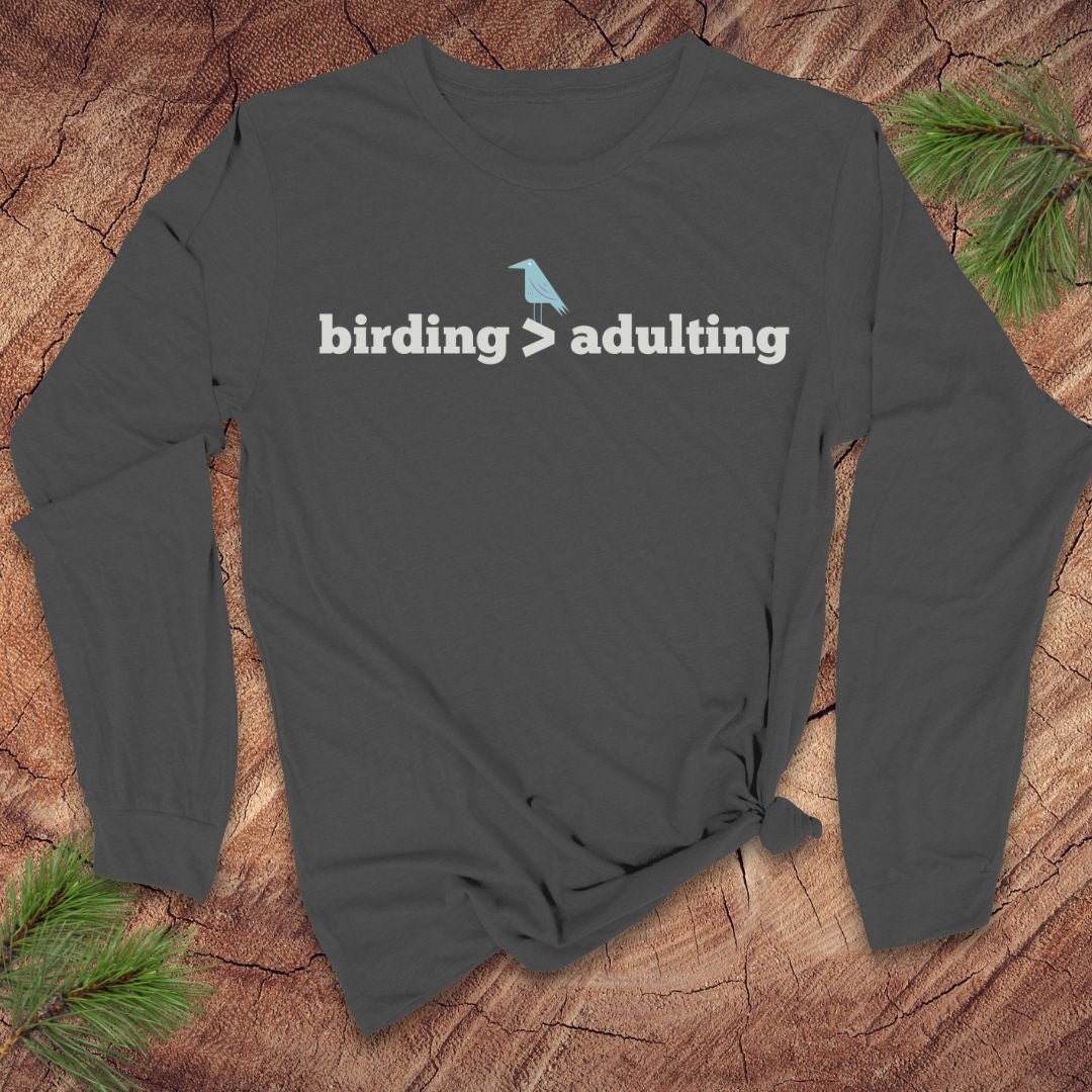 Dark gray long-sleeve shirt with 'birding > adulting' text on a wooden surface with pine branches.
