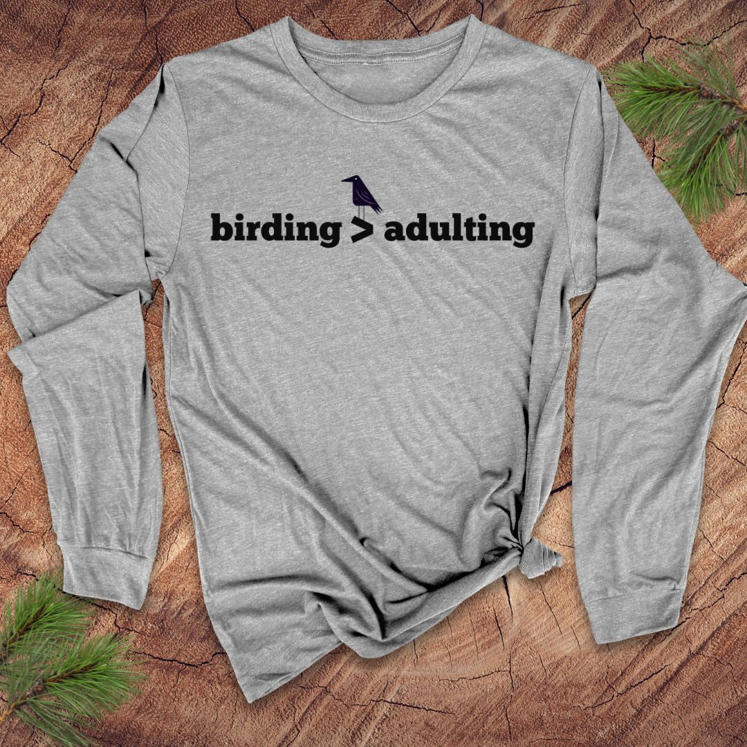 Gray long-sleeve shirt with 'birding > adulting' text on a wooden surface with pine branches.