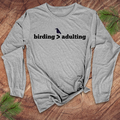 Gray long-sleeve shirt with 'birding > adulting' text on a wooden surface with pine branches.