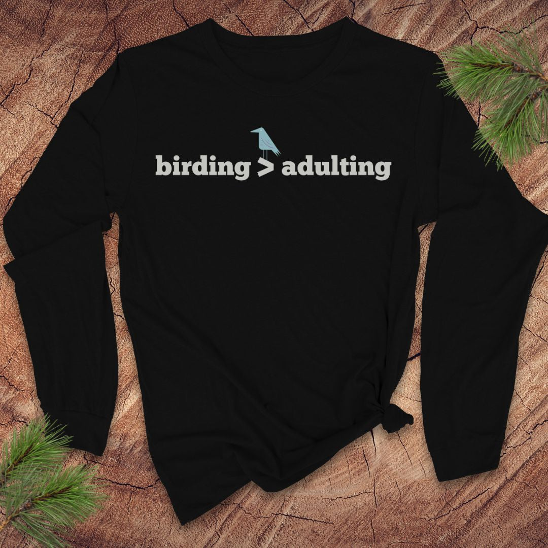 Black long-sleeve tshirt with 'birding > adulting' text on a wooden surface with pine branches.