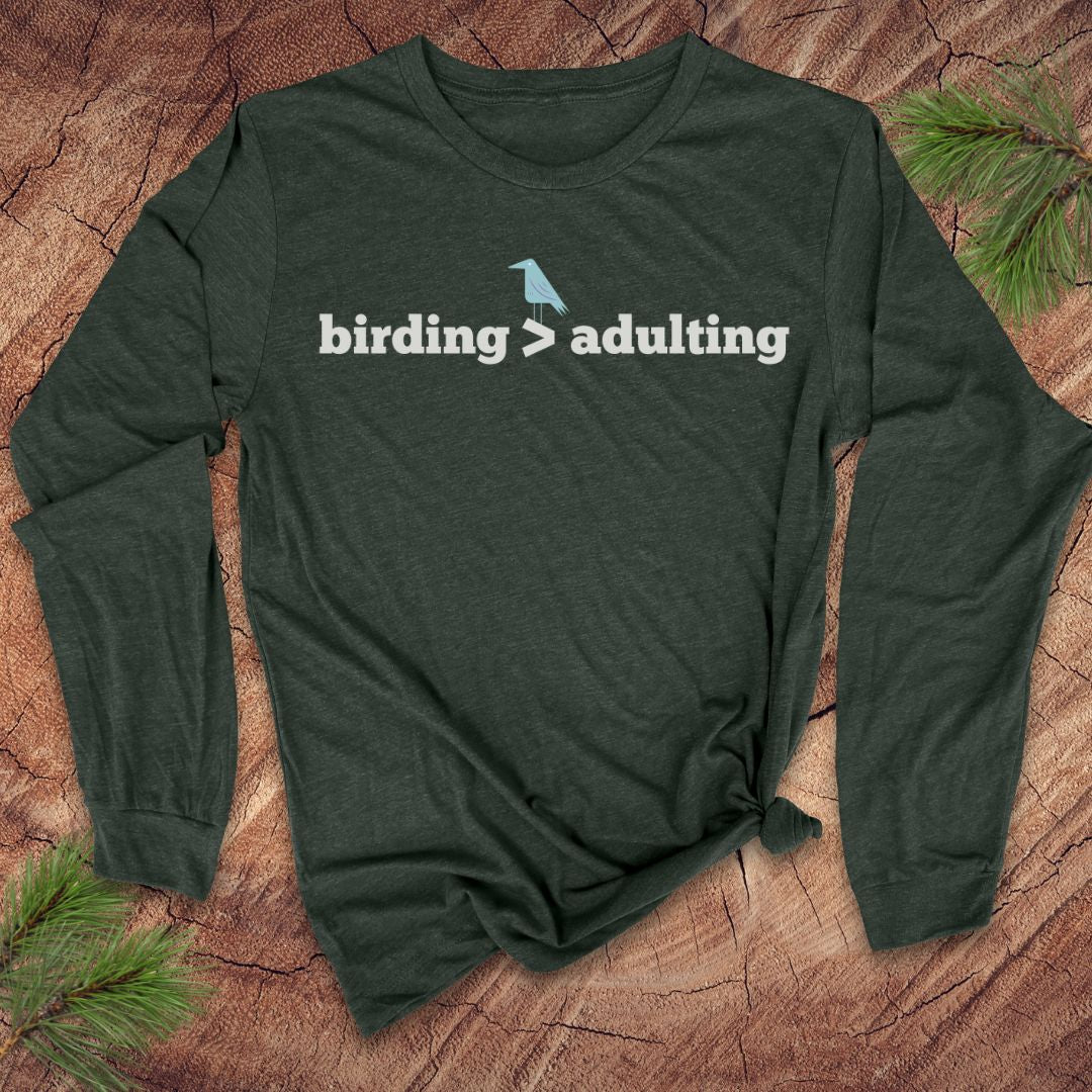 Green long-sleeve shirt with 'birding > adulting' text on a wooden surface with pine branches.
