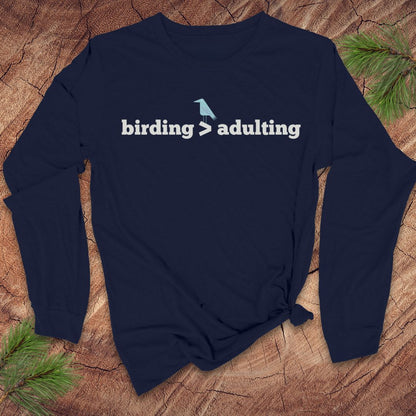 Navy blue long-sleeve tshirt with 'birding > adulting' text on a wooden surface with pine branches.