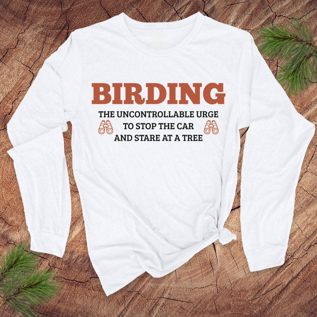 White long sleeve tshirt with funny 'BIRDING' text on a wooden surface with pine branches.