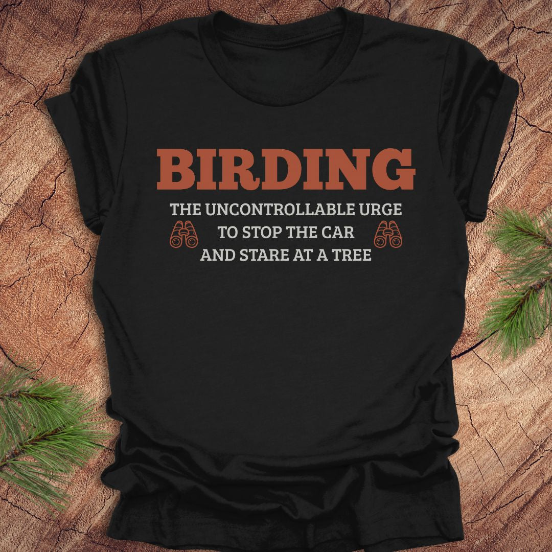 Black t-shirt with birding-themed text on a wooden surface with pine branches.