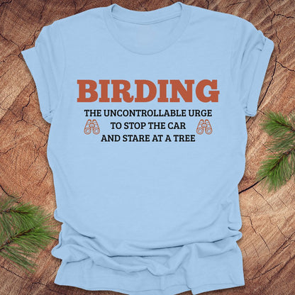 Light blue t-shirt with birding-themed text on a wooden surface with pine branches.