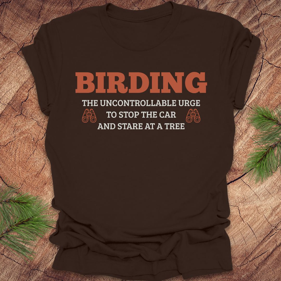Brown t-shirt with 'BIRDING' text on a wooden surface with pine branches.