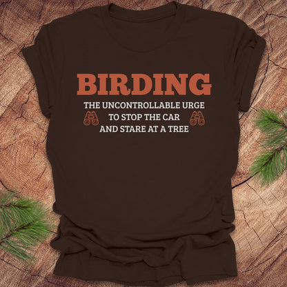 Brown t-shirt with 'BIRDING' text on a wooden surface with pine branches.