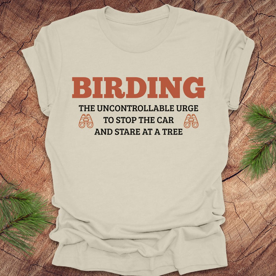 Beige t-shirt with 'BIRDING' text on a wooden surface with pine branches.