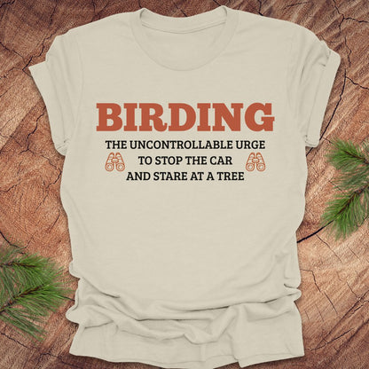 Beige t-shirt with 'BIRDING' text on a wooden surface with pine branches.