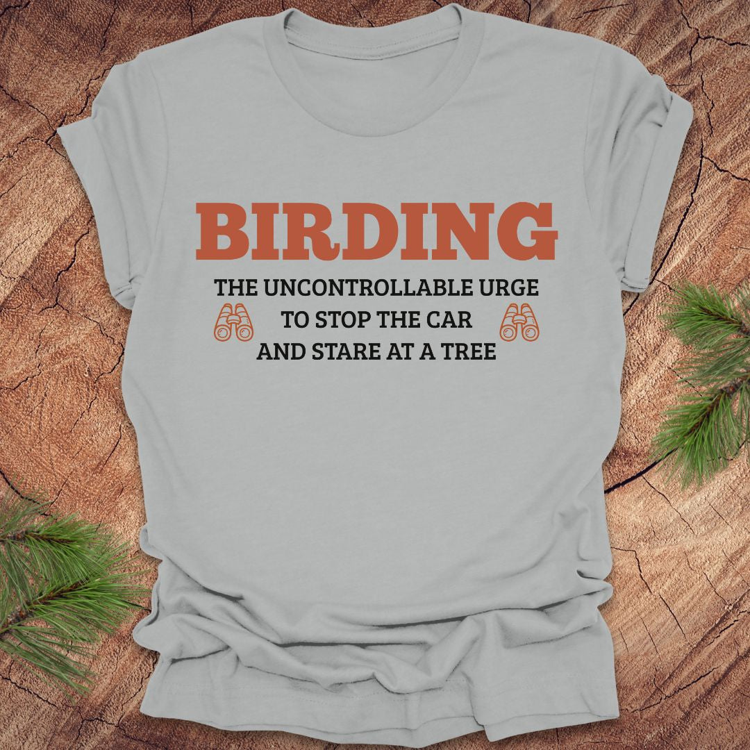 Gray t-shirt with birding-themed text on a wooden surface with pine branches.