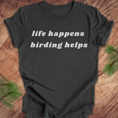 A dark charcoal gray t-shirt with the phrase 'life happens birding helps' printed in white serif text across the chest, laid flat on a rustic wood background with pine branches in the corners.