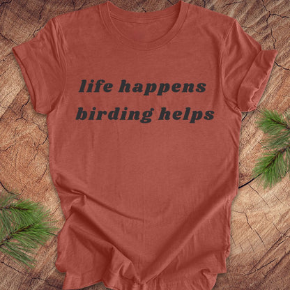 A terracotta-colored short-sleeve t-shirt with the text 'life happens birding helps' printed in black across the chest, laid flat on a rustic wood slice background with pine needles in the corners.