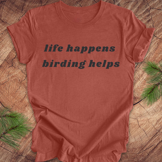 A terracotta-colored short-sleeve t-shirt with the text 'life happens birding helps' printed in black across the chest, laid flat on a rustic wood slice background with pine needles in the corners.