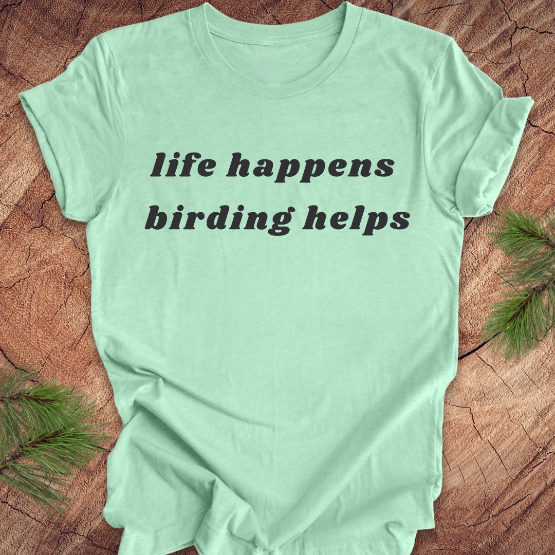 A mint green short-sleeve t-shirt with the text 'life happens birding helps' printed in black serif font across the chest, laid flat on a rustic wood surface with pine branches in the corners.