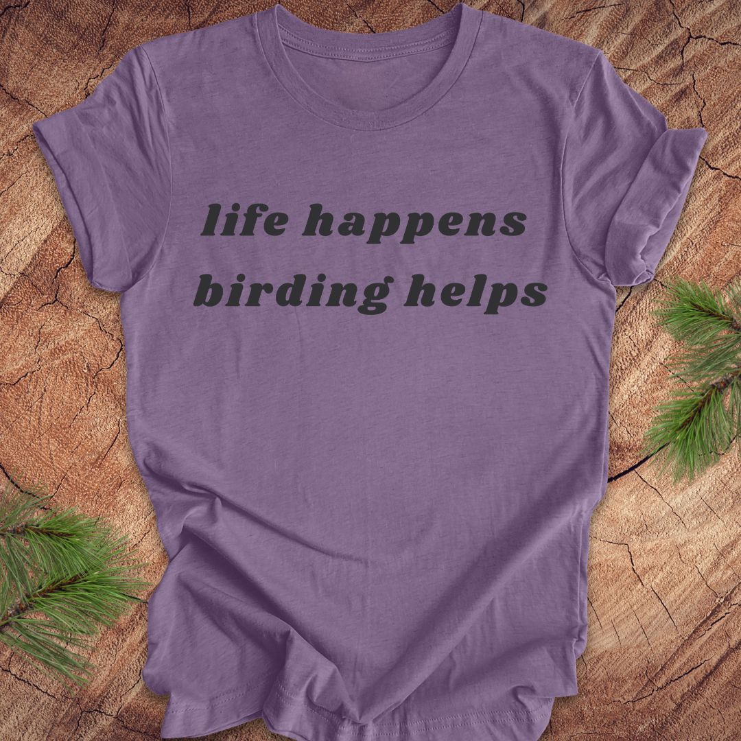 A purple short-sleeve crewneck t-shirt with the text 'life happens birding helps' printed in black across the chest, laid flat on a rustic wood background with pine branches in the corners.