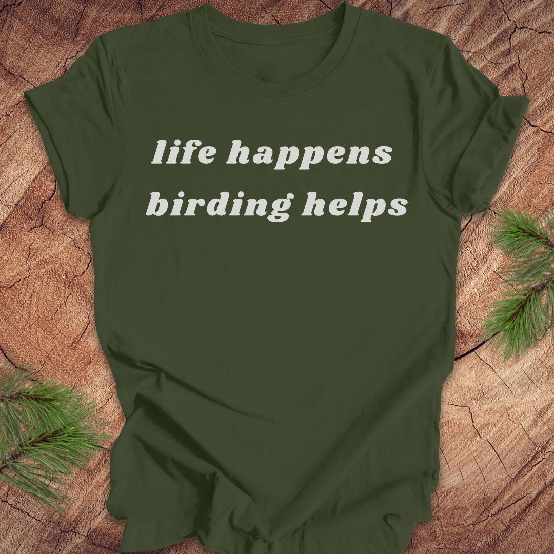 An olive green short-sleeve t-shirt with the phrase 'life happens birding helps' printed in white serif text across the chest, laid flat on a rustic wood slice background with pine branches in the corners.