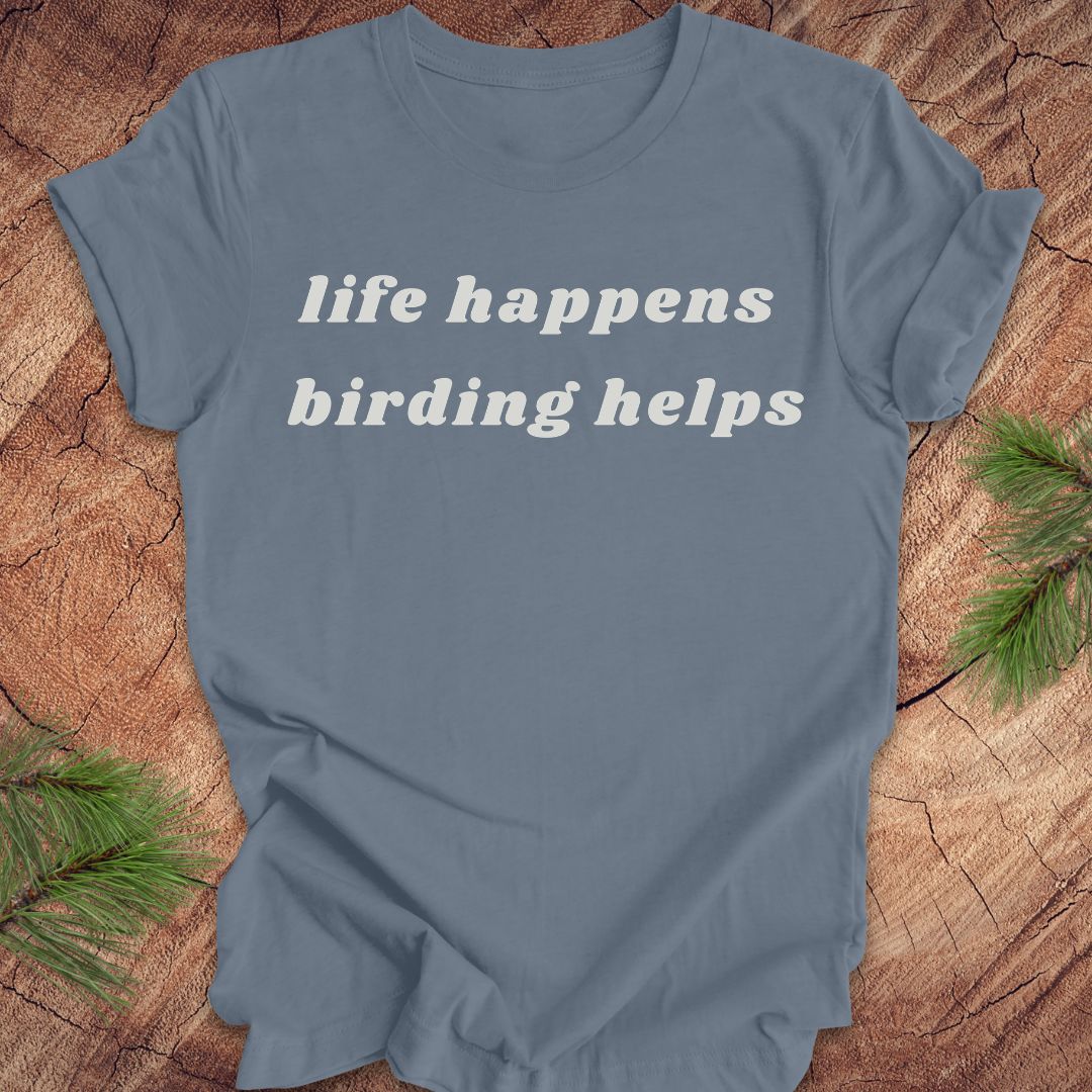 A slate blue crewneck t-shirt with the phrase 'life happens birding helps' printed in white serif text across the chest, laid flat on a rustic wood slice background with pine needles in the corners.