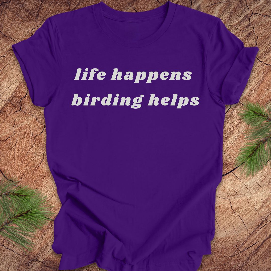 A purple short-sleeve t-shirt with the phrase 'life happens birding helps' printed in white serif text across the chest, laid flat on a rustic wood surface with pine branches in the corners.