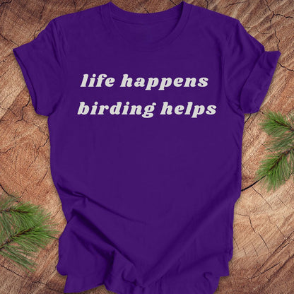 A purple short-sleeve t-shirt with the phrase 'life happens birding helps' printed in white serif text across the chest, laid flat on a rustic wood surface with pine branches in the corners.