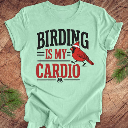 Light green t-shirt with 'BIRDING IS MY CARDIO' text and a cardinal graphic on a wooden surface.