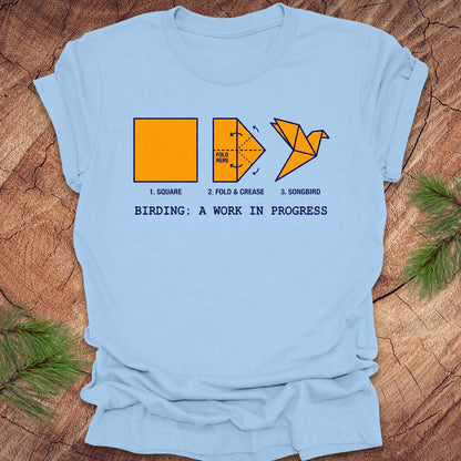 Light blue t-shirt with origami bird design and text on a wooden surface.