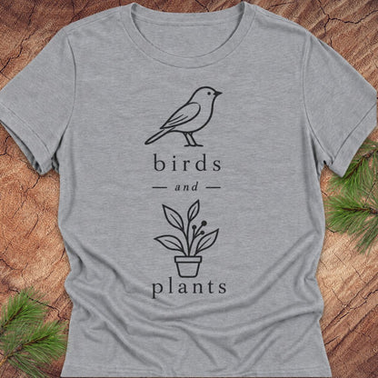 A heather gray short-sleeve t-shirt with a black line art graphic of a bird, a potted plant, and the words 'birds and plants'. The shirt is laid flat on a rustic wood background with pine needles in the corners.
