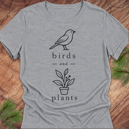 A heather gray short-sleeve t-shirt with a black line art graphic of a bird, a potted plant, and the words 'birds and plants'. The shirt is laid flat on a rustic wood background with pine needles in the corners.