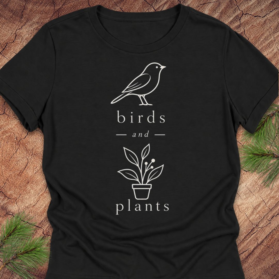 A black short-sleeve crewneck t-shirt with a white line art graphic of a bird, the text 'birds and plants', and a potted plant, laid flat on a rustic wood background with pine needles.