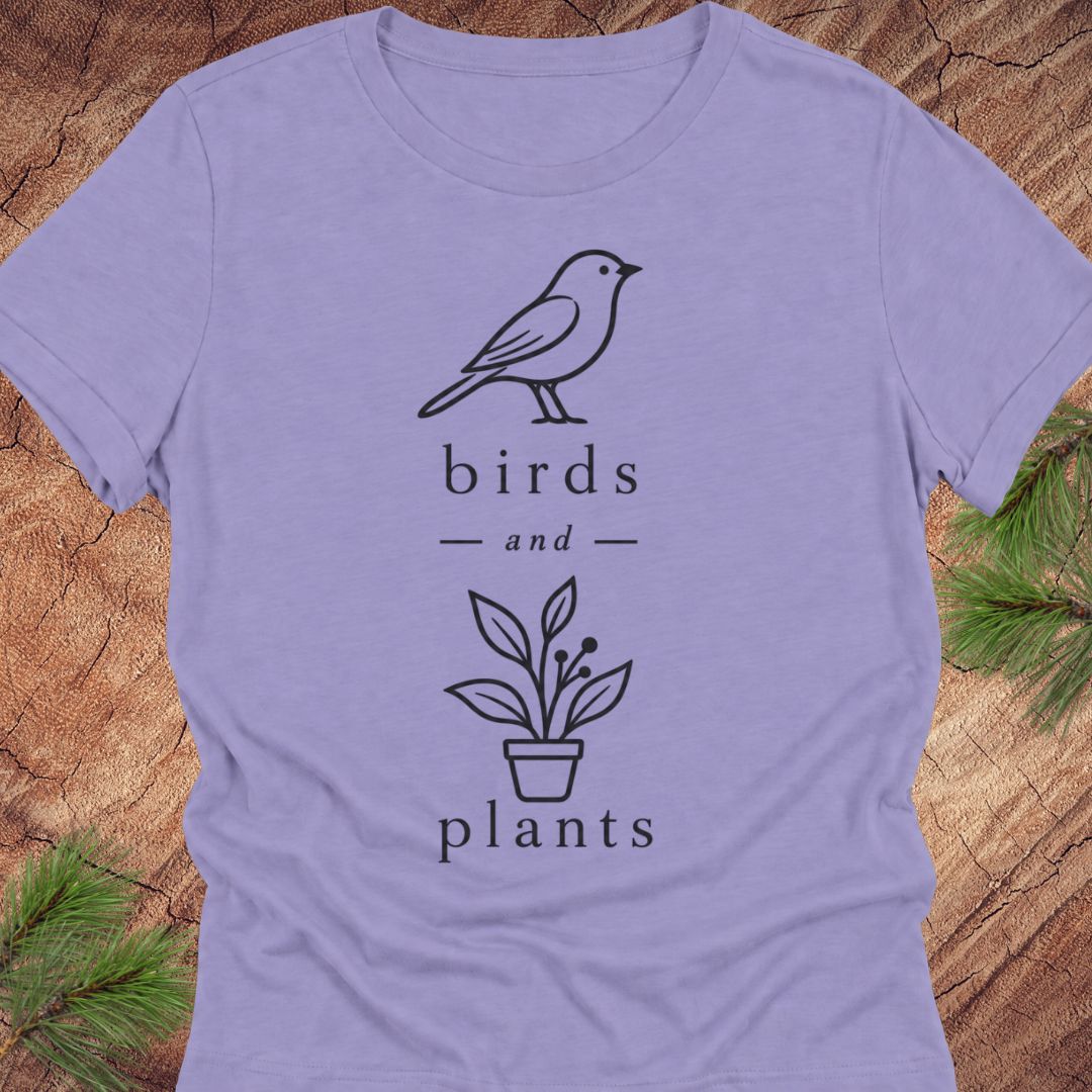 A lavender short-sleeve crewneck t-shirt with a black graphic print of a bird, the words 'birds and plants', and a potted plant, laid flat on a rustic wood background with pine needles.