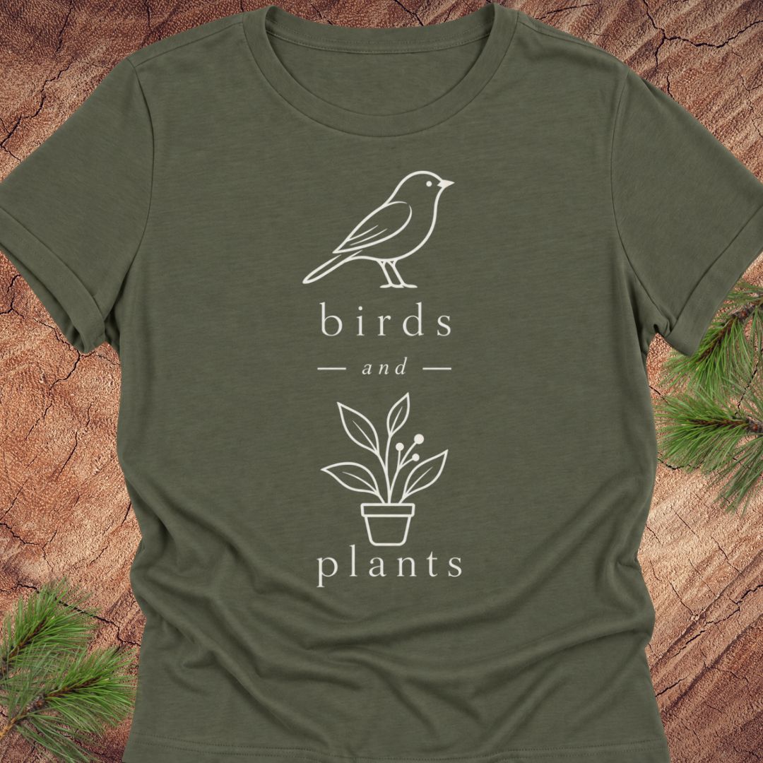 An olive green short-sleeve t-shirt with a white line art graphic of a bird and a potted plant, with the words 'birds and plants' printed on the front. The shirt is laid flat on a rustic wood background with pine branches in the corners.