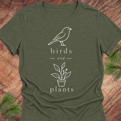 An olive green short-sleeve t-shirt with a white line art graphic of a bird and a potted plant, with the words 'birds and plants' printed on the front. The shirt is laid flat on a rustic wood background with pine branches in the corners.