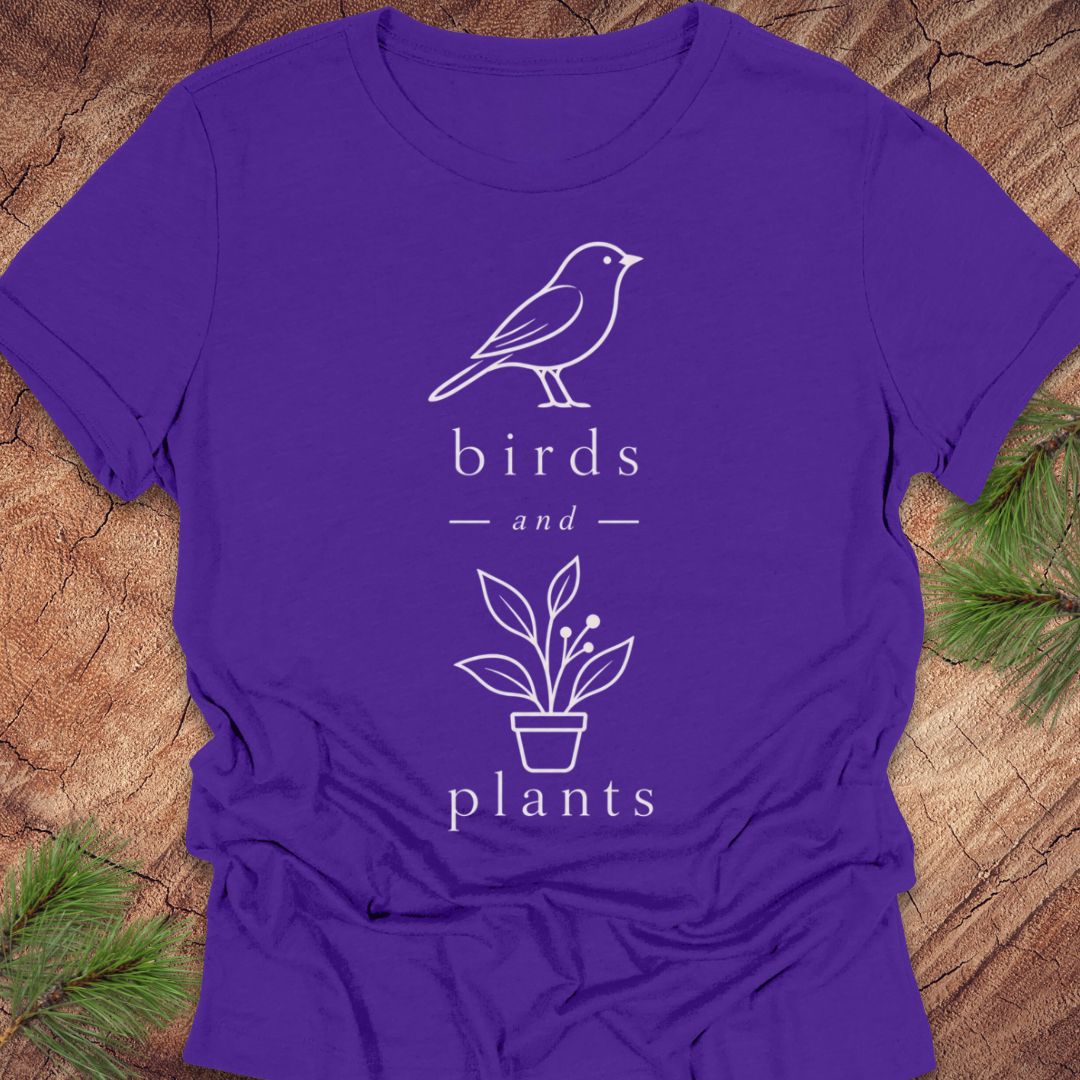 A purple short-sleeve t-shirt with a white line art graphic of a bird, the words 'birds and', a potted plant, and the word 'plants', laid flat on a rustic wood background with pine branches in the corners.