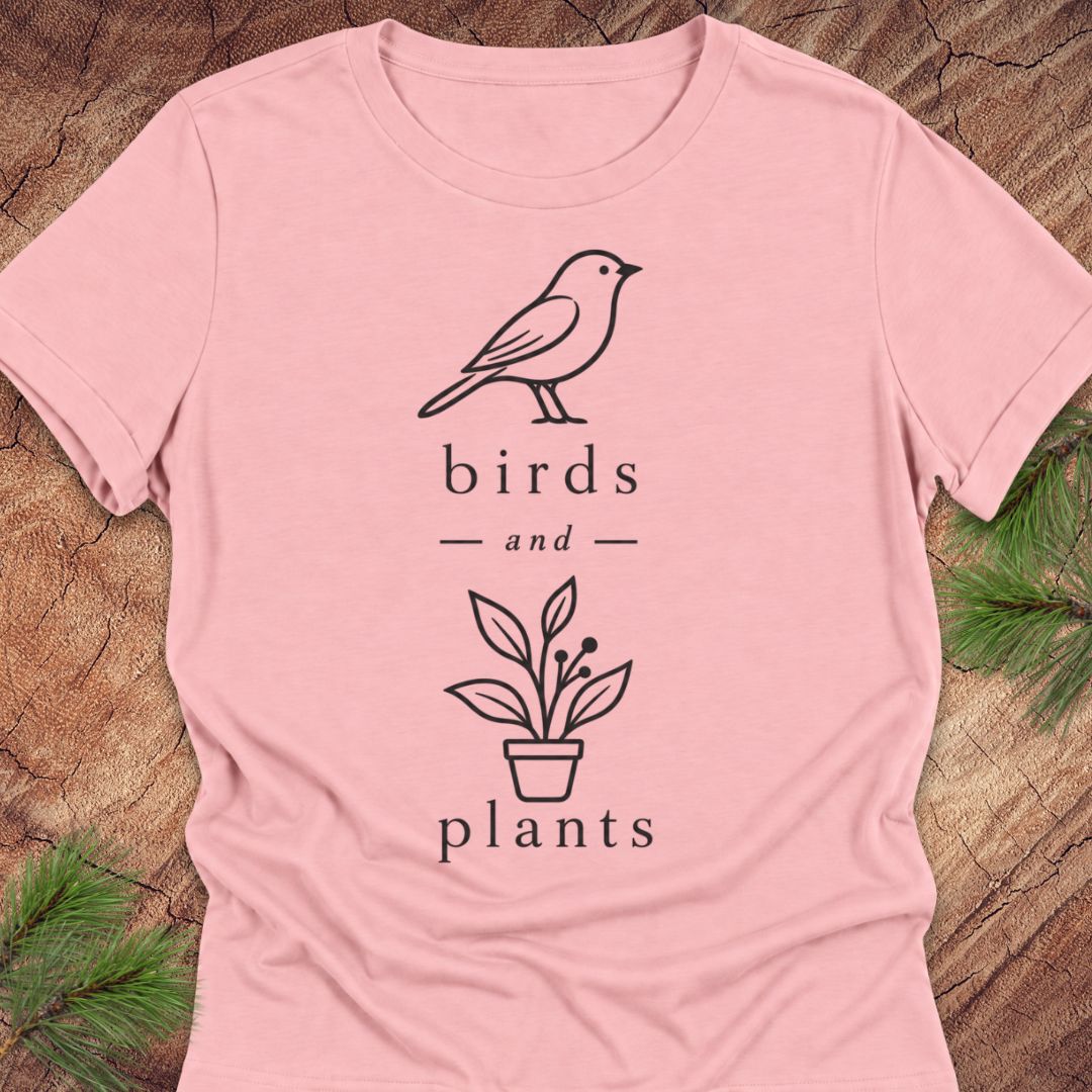 A light pink short-sleeve t-shirt with a black line art graphic of a bird and a potted plant, with the words 'birds and plants' printed in a serif font. The t-shirt is laid flat on a rustic wood background with green pine branches in the corners.